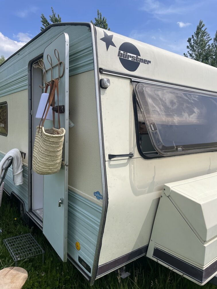 Retro Bűrstner City caravan with a light blue exterior, door open, and a woven bag hanging, set against a clear sky.